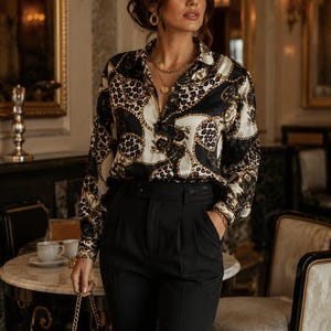 Könnte beinhalten: Eine Frau trägt ein Seidenhemd mit einem Muster aus schwarzen, weißen und goldenen Ketten und Leopardenmuster. Dazu eine schwarze Hose und Goldschmuck. Das Outfit wirkt elegant und anspruchsvoll, passend für formelle Anlässe oder einen stilvollen Auftritt.