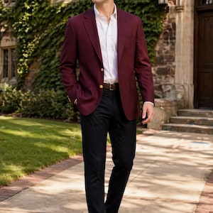 May include: A burgundy blazer with gold buttons, paired with a white dress shirt and black trousers. The outfit is completed with a brown leather belt and brown leather loafers. The man is walking on a brick and concrete walkway.
