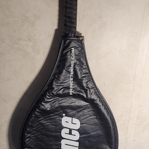 May include: Black tennis racket with a black grip. The racket has the brand name "Prince" in white lettering. The website address "princetennis.com" is also visible on the racket.