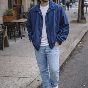 May include: A person wearing a navy blue jacket, white t-shirt, light blue jeans, and white sneakers. The person is standing on a city sidewalk. The jacket has a collared neckline and button closure.