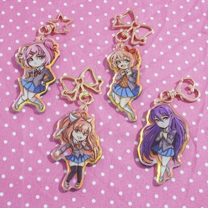 Puede incluir: Cuatro llaveros de personajes de anime coloridos con detalles dorados. Cada llavero presenta un personaje diferente con cabello rosa, azul, morado o rubio. Los llaveros están sobre un fondo de lunares rosa y blanco.