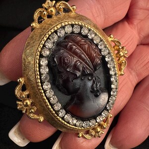 May include: Oval gold-toned cameo brooch featuring a dark profile portrait within a frame of clear crystals. The cameo is set in a textured gold frame with ornate floral accents. A classic piece of jewelry.