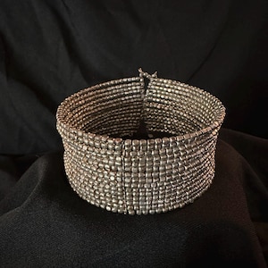 May include: A wide silver-toned bracelet composed of numerous small, rectangular beads. The bracelet has a cuff design and appears to be made of metal. The beads are tightly woven together, creating a textured surface. The bracelet is displayed against a dark background.