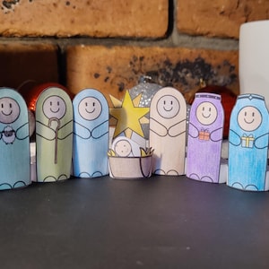 May include: A handmade Christmas nativity scene with paper cutouts. The figures include an angel, shepherds, the baby Jesus, and the three wise men. Each figure has a smiling face and is colored with markers. The background includes a brick wall and a Christmas card.