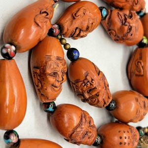 May include: Close-up of a necklace featuring carved orange beads with detailed faces and other shapes. The beads are strung together with smaller, colorful accent beads. The necklace has a vintage or antique aesthetic.