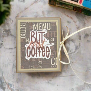 May include: A small brown box with a beige ribbon tied around it. The box has a brown and beige patterned cover with the words "But first coffee" printed in brown on a white background.