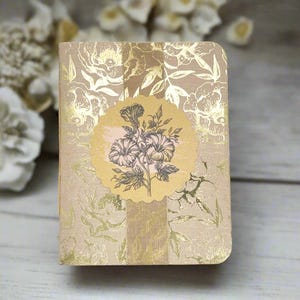 May include: A small, closed notebook with a floral design. The cover is a light beige color with a gold floral pattern. A round, light yellow sticker with a floral illustration is centered on the cover. A vertical gold stripe runs down the center of the notebook.