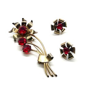 May include: A vintage jewelry set featuring a gold-toned brooch and matching earrings. The brooch is shaped like a floral bouquet with three red gemstone accents and a bow. The earrings are small, gold-toned, and feature a single red gemstone.
