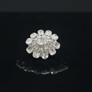 May include: A clear, flower-shaped rhinestone button with a central rectangular stone surrounded by smaller, faceted stones. The button is set against a black background, creating a striking contrast. This button is ideal for embellishing clothing or accessories.