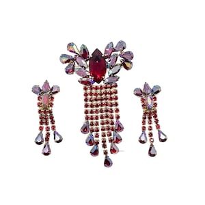 May include: A vintage jewelry set featuring a brooch and matching earrings. The set is adorned with ruby red and iridescent crystals. The brooch has a large central stone and a cascading fringe of smaller crystals. The earrings have a similar design.