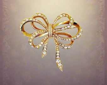 May include: A gold-toned bow brooch, encrusted with sparkling clear crystals. The brooch features a classic bow design with looped ribbons and dangling ends. The crystals are set in a pave style, adding brilliance to the jewelry.