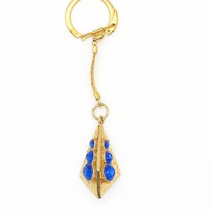 May include: A gold-colored keychain with a diamond-shaped pendant. The pendant features a textured surface and is adorned with several blue, gem-like accents. The keychain includes a chain and a clasp for attaching to keys or other items.