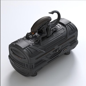 May include: A black, cylindrical container with a detailed, textured surface. A sculpted, black alien figure with a curved tail and detailed features is perched on top. The container has a hinged design and appears to be a storage box or case.