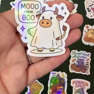 Könnte beinhalten: Ein holografischer Aufkleber mit einer Kuh in einem Geisterkostüm mit dem Text "Mooo I mean Boo" in einer Sprechblase. Weitere Aufkleber mit Herbst- und Halloween-Themen sind sichtbar.