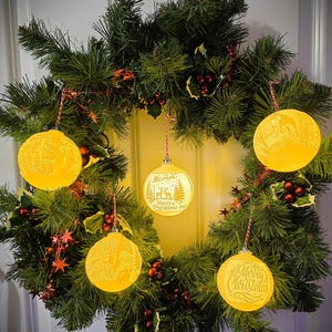 May include: A Christmas wreath with green pine needles, red berries, and star-shaped ornaments. Five illuminated yellow Christmas ornaments are suspended from the wreath, each with a unique holiday design and text that reads "Merry Christmas".