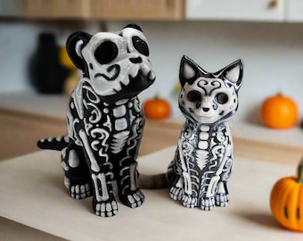 May include: Two small, black and white skeletal animal figurines. The larger figurine is a dog, and the smaller figurine is a cat. Both figurines have intricate, detailed designs.