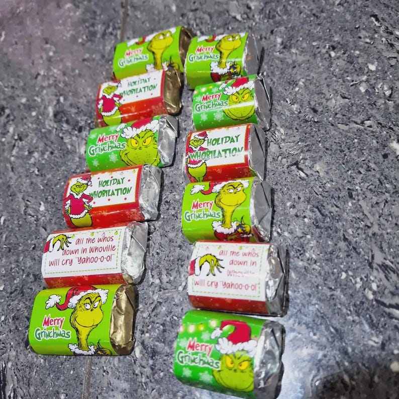 K&ouml;nnte beinhalten: Eine Sammlung einzeln verpackter Schokoriegel mit festlicher Verpackung. Die Verpackungen zeigen den Grinch-Charakter in verschiedenen Posen, zusammen mit dem Text "Merry Grinchmas" und "Holiday Whobilation". Einige Verpackungen enthalten auch den Satz "all the whos down in Whoville will cry Yahoo-o-o".