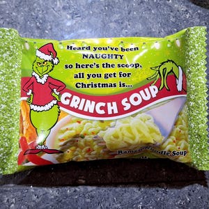 Könnte beinhalten: Eine Packung Grinch Soup Ramen-Nudeln. Die Verpackung ist grün mit einem Cartoon-Grinch, der einen Weihnachtsmannhut und einen roten Anzug trägt. Auf der Verpackung steht "Heard you've been NAUGHTY... GRINCH SOUP". Die Ränder der Verpackung sind mit einem grün-weißen Muster verziert.