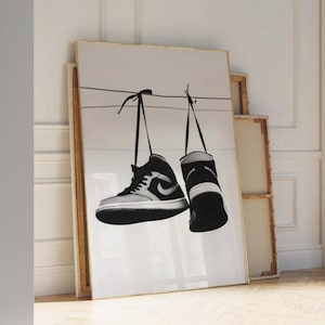 Può includere: Stampa artistica in bianco e nero di due scarpe da ginnastica appese a un filo. Le scarpe alte hanno un design classico con un logo a virgola. L'opera d'arte è incorniciata in una cornice dorata.