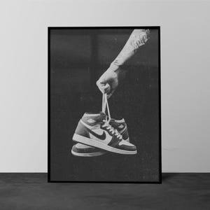 Può includere: Stampa incorniciata in bianco e nero con un paio di sneakers alte. Le scarpe sono tenute per i lacci, con una mano visibile. Le scarpe hanno una base bianca, dettagli neri e un iconico logo Nike.