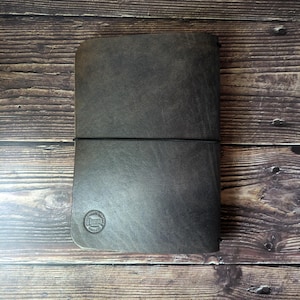 Puede incluir: Una funda de diario de cuero marrón oscuro con una textura suave y mate y esquinas redondeadas. Una fina banda elástica negra envuelve horizontalmente el centro para asegurar la funda. Un pequeño logotipo circular está grabado en la esquina inferior izquierda.