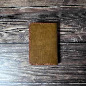 May include: A brown leather wallet with red stitching around the edges. The wallet is rectangular and appears to be closed. The background is a dark wood surface.