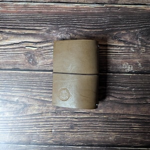 Peut inclure: Un journal en cuir beige avec une fermeture élastique marron foncé. Le journal a un emblème circulaire en relief sur la couverture avant. Le journal est rectangulaire et semble fermé.