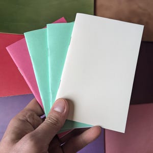 Pode incluir: Uma mão segura uma pilha de pequenos cadernos com capas brancas, rosa e verde menta. Os cadernos estão em branco e têm encadernação costurada. O fundo apresenta outros papéis coloridos.