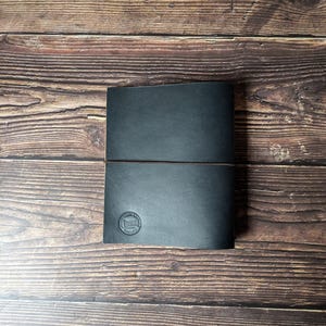 May include: A rectangular black leather journal featuring a thin horizontal elastic band closure. The cover displays a circular embossed logo in the bottom left corner with the text "DEPARTURE LEATHER ENGLAND" encircling a small graphic icon.