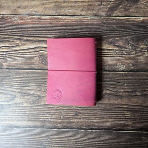 May include: A magenta leather journal with a debossed emblem on the front cover. The journal is closed with an elastic band. The journal is on a wooden surface.