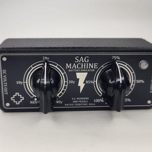 May include: Black "SAG MACHINE" battery emulator with two large knobs and white text. The device has a lightning bolt graphic and reads "E.G. Wonders BMF Pedals Native Territory, Okla."