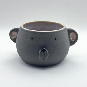 May include: A dark grey ceramic bowl shaped like a bear head with two protruding ears, two small dot eyes, and a raised snout featuring a black nose. The interior of the bowl has a smooth, light purple glaze, while the exterior has a matte finish.