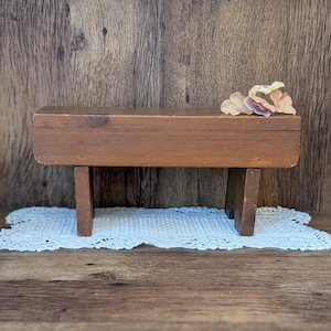 May include: A small, rectangular wooden bench with four legs. The bench is made of stained wood and has a natural finish. A decorative floral arrangement sits on top of the bench. The bench is placed on a white, lace doily.