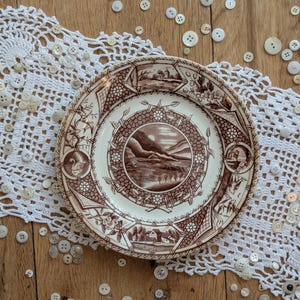 May include: A round ceramic plate featuring a brown and white transferware pattern with a central landscape scene, surrounded by smaller vignettes of swans and buildings, resting on a white crocheted lace doily scattered with white buttons.