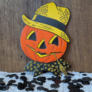 May include: A vintage Halloween decoration featuring a smiling orange pumpkin wearing a yellow hat with a black brim. The pumpkin has black eyes and a mouth, and a piece of wheat in its mouth. Black buttons are scattered on a white lace surface.