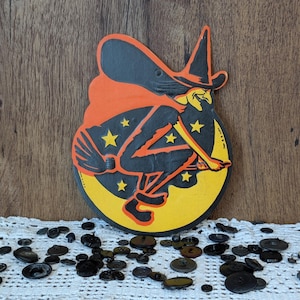 May include: A vintage Halloween decoration featuring a witch riding a broom against a yellow moon with stars. The witch is dressed in black and orange, with a pointed hat. The decoration is on a wooden surface.