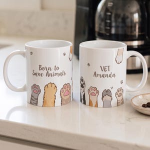 Puede incluir: Dos tazas de cerámica blanca con ilustraciones coloridas de varias patas de animales en el borde inferior. La taza izquierda muestra el texto Born to Save Animals, mientras que la derecha presenta el texto VET Amanda en letra cursiva gris.