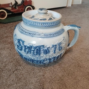 May include: A white and blue ceramic crock with a lid. The crock has a handle and is decorated with blue floral patterns and the words "Spirit of 76". The lid has a knob and a blue floral design.