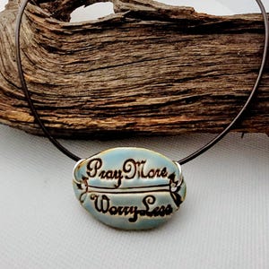 May include: A necklace with a light blue oval pendant that reads "Pray More, Worry Less" in script. The pendant is on a black cord. The background is a piece of driftwood.