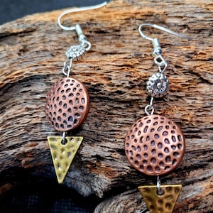May include: A pair of dangle earrings with a copper and gold geometric design. The earrings feature a round copper disc with a textured surface, a gold triangle, and a silver flower charm.