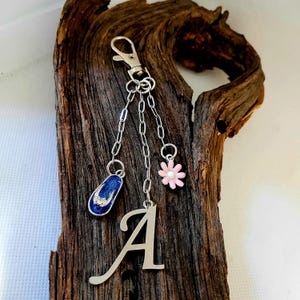 May include: Silver-toned keychain with a blue flip-flop charm, a pink flower charm, and a silver letter "A" charm. The flip-flop has a blue enamel base with a sparkly design. The flower has a white center.
