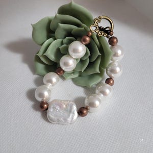 May include: A pearl bracelet with a square, iridescent center bead and round white pearls. The bracelet has bronze-colored beads and a decorative clasp. The bracelet is displayed on a green succulent.