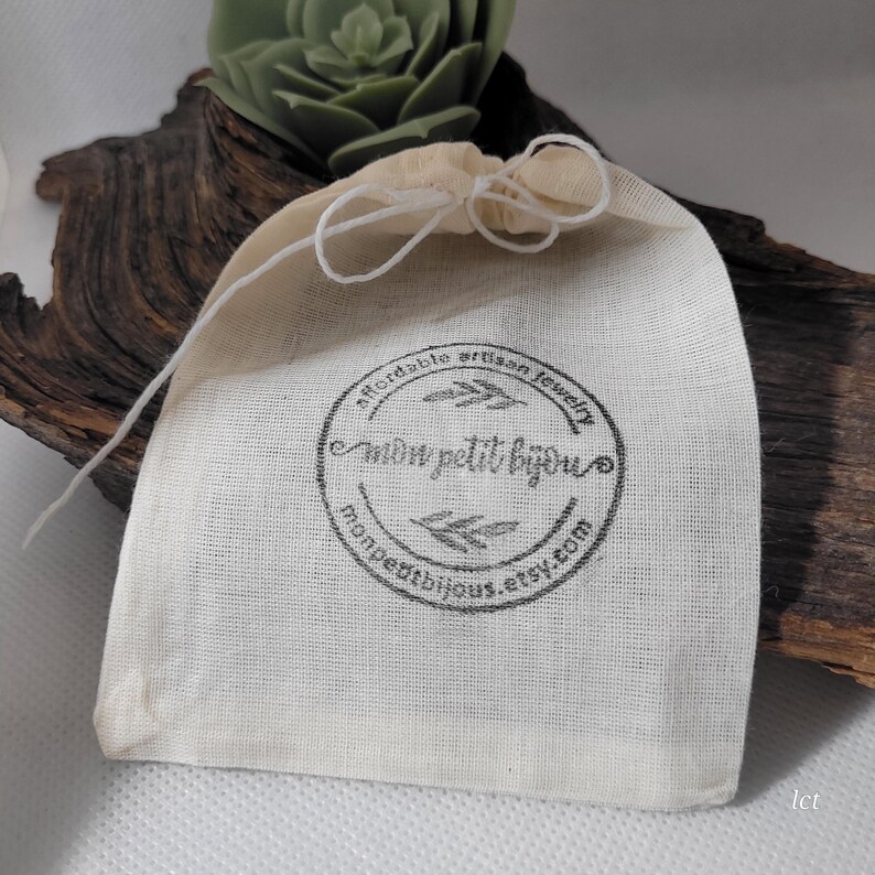 May include: A small, off-white drawstring bag with the text "mon petit bijou" in a circular design. The bag is made of a natural fabric and is tied with a white string. The background includes a succulent and a piece of driftwood.