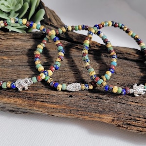 May include: Three beaded bracelets with colorful beads in shades of blue, red, yellow, and green. Each bracelet features a different silver charm: an elephant, a snake, and a turtle. The bracelets are displayed on a piece of driftwood.