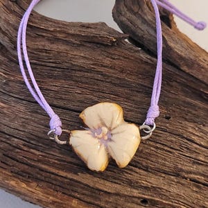 May include: A handmade bracelet featuring a cream-colored flower charm with a purple cord. The flower charm is detailed with brown accents and is attached to the cord with silver-colored rings. The bracelet is displayed on a piece of driftwood.