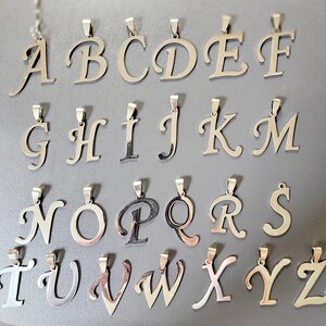 May include: Silver-toned alphabet pendants, each with a loop for a chain. The letters A through Z are displayed in a cursive font. The pendants are arranged on a gray surface, showcasing the complete alphabet.
