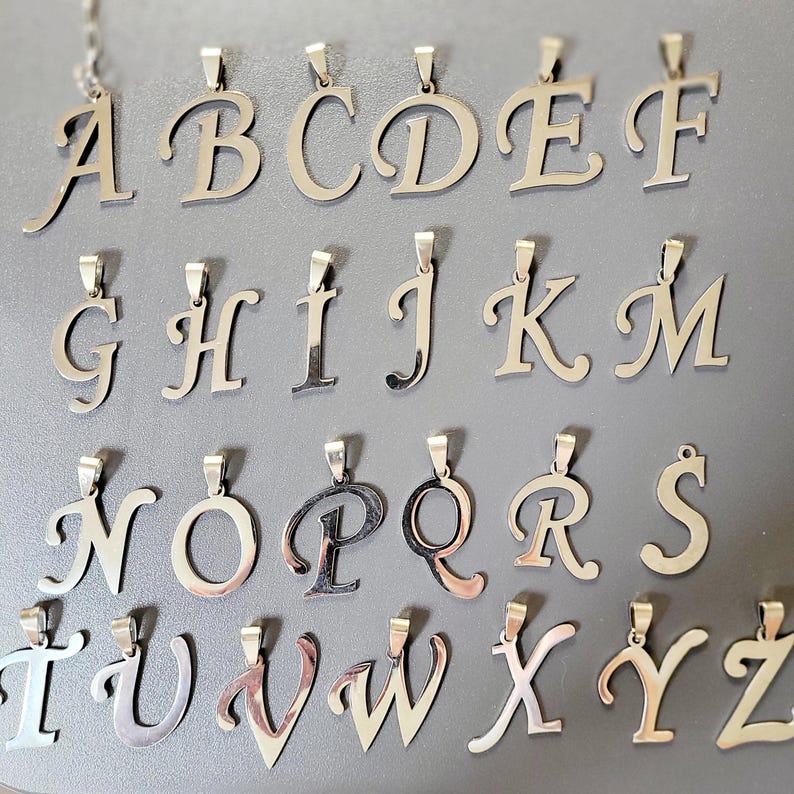 May include: Silver-toned alphabet pendants, each with a loop for a chain. The letters A through Z are displayed in a cursive font. The pendants are arranged on a gray surface, showcasing the complete alphabet.