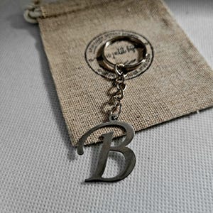 May include: A silver-colored metal keychain featuring the letter "B" in a stylized font. The keychain is attached to a chain and a keyring. It rests on a small, beige drawstring bag with a circular stamp design.