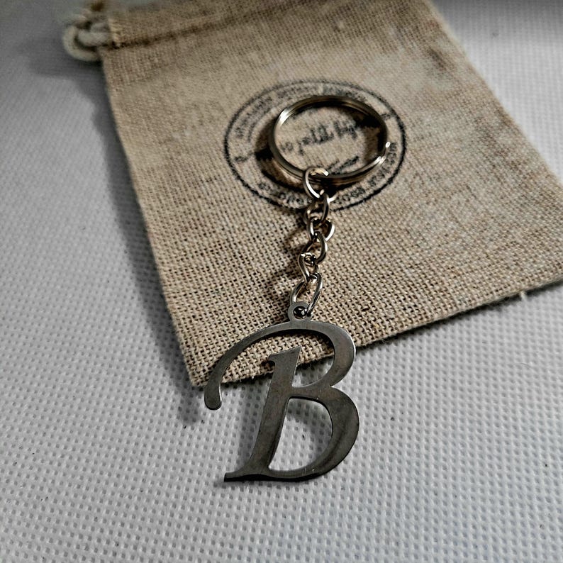 May include: A silver-colored metal keychain featuring the letter "B" in a stylized font. The keychain is attached to a chain and a keyring. It rests on a small, beige drawstring bag with a circular stamp design.