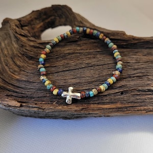May include: A beaded bracelet with a silver cross charm. The bracelet features small, colorful beads in shades of yellow, blue, red, and brown. The cross is centered on the bracelet, which is displayed on a piece of driftwood.